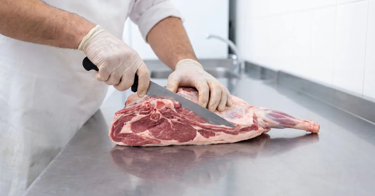 Close-up of gloved hands cutting a fresh leg of lamb on a steel counter